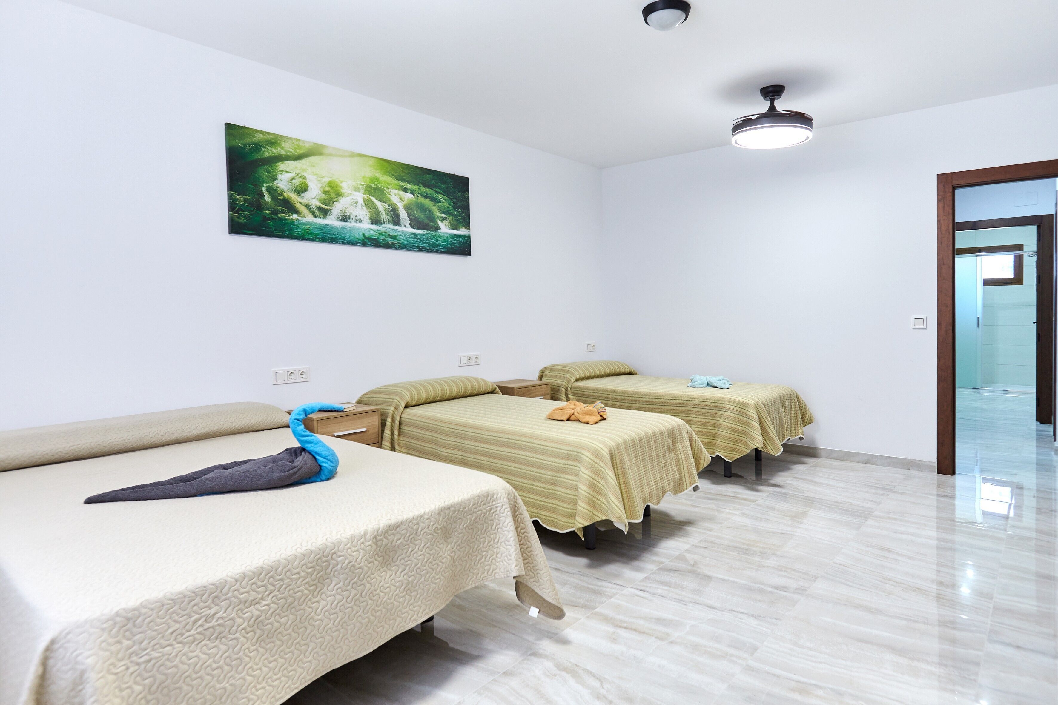 A hotel room with three beds, a ceiling fan, a wall-mounted picture, and a door leading to another room.