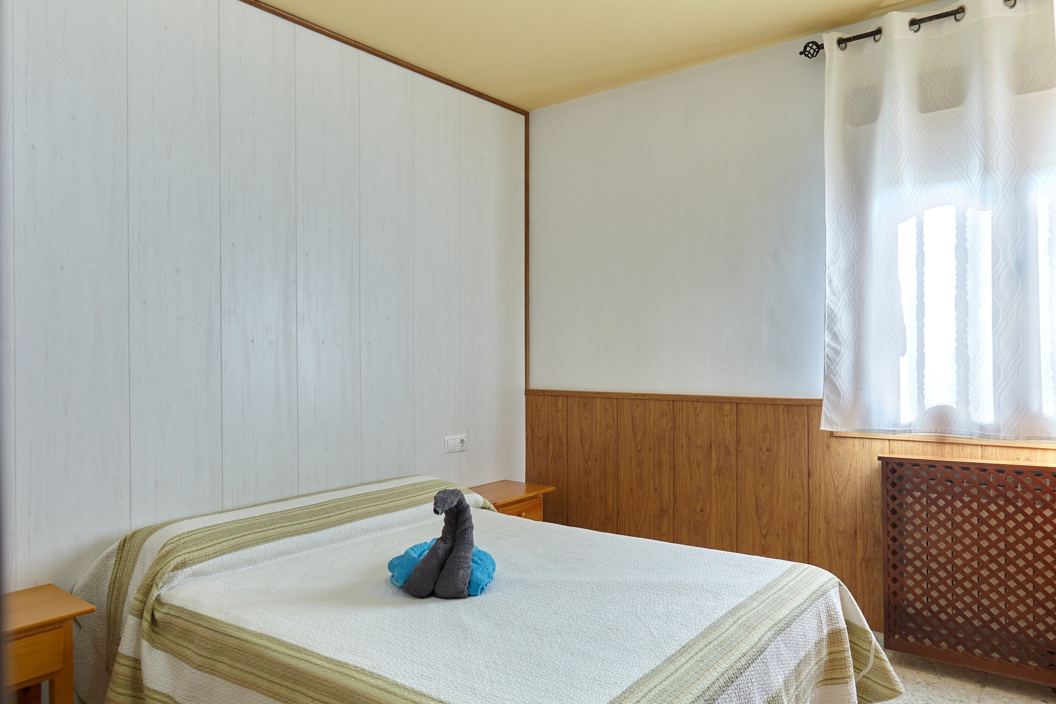 Un dormitorio con cama, mesita de noche, pared de paneles de madera, ventana con cortina y radiador.