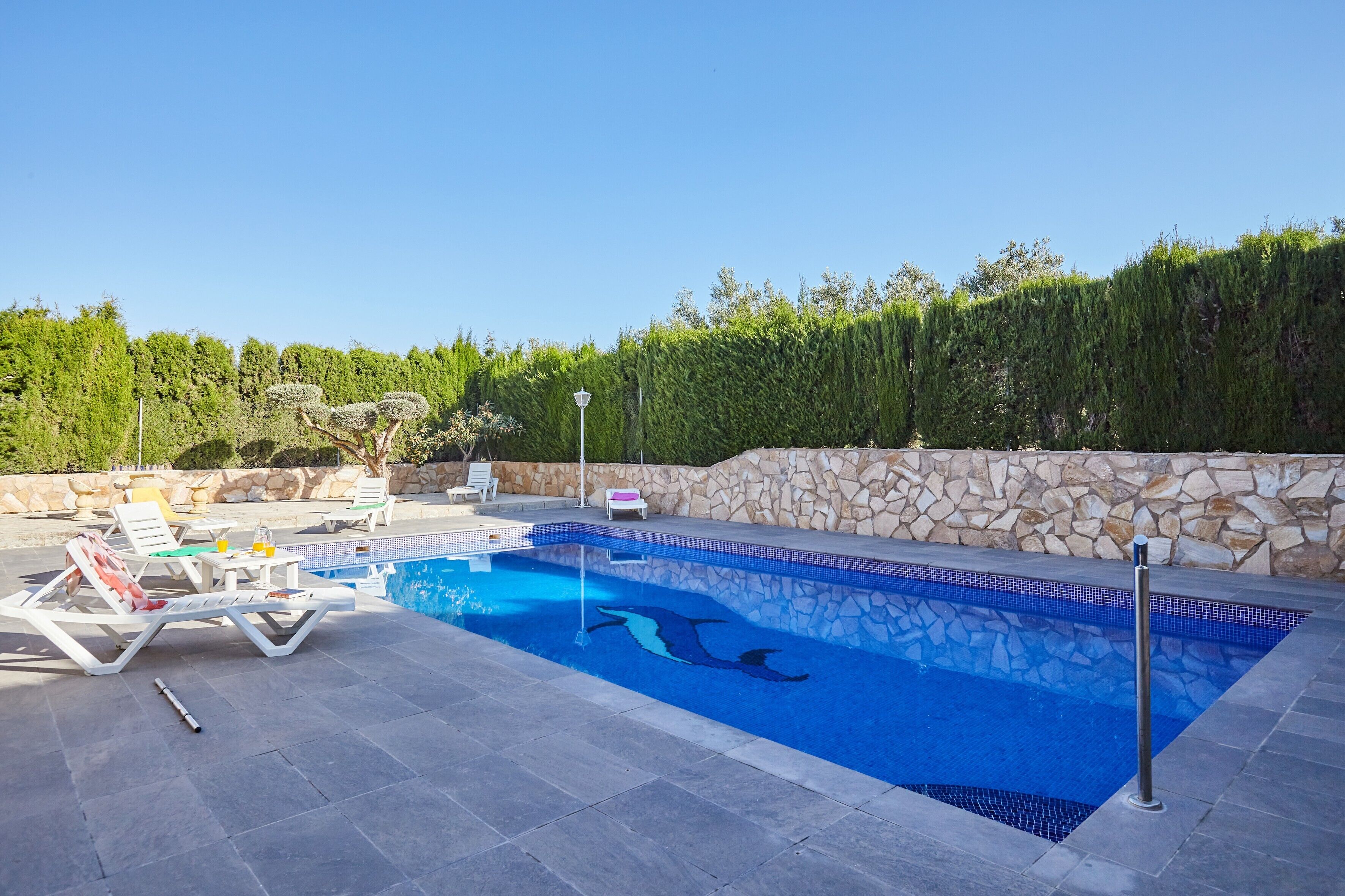 Una piscina con sillas de descanso, un muro de piedra y un seto al fondo.