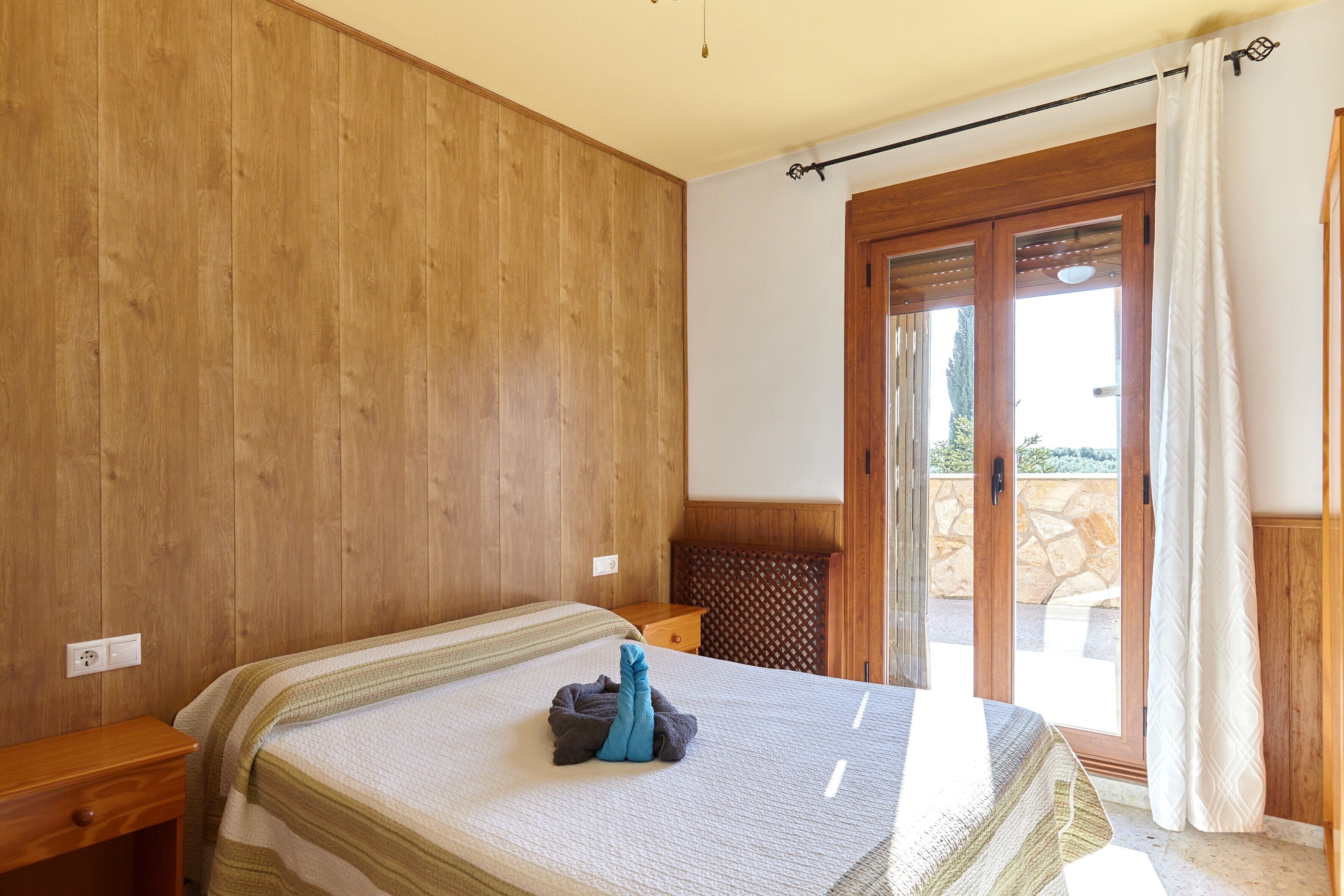 Een slaapkamer met een houten wand, een bed met een gestreept dekbed, een nachtkastje en een schuifdeur naar een buitenruimte.