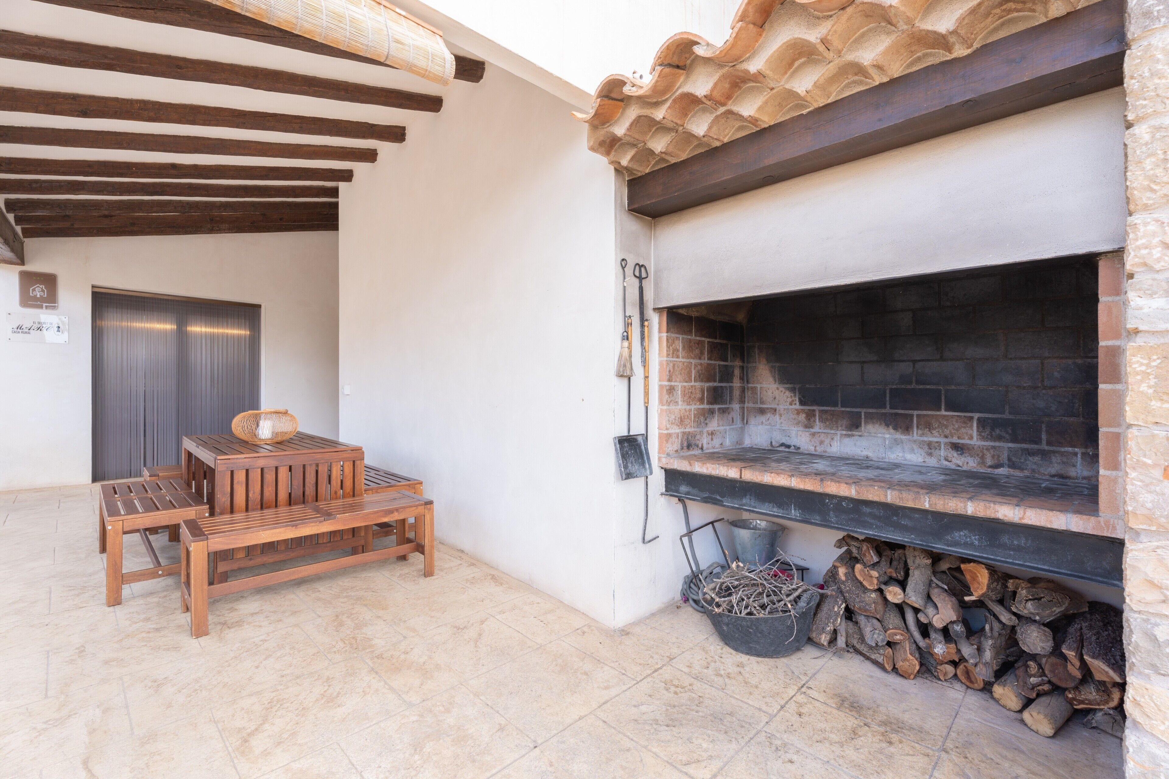 A patio with a wooden table and benches, a stone fireplace, and a door in the background.