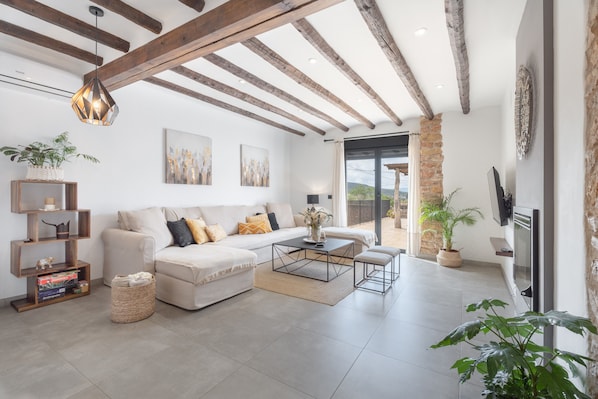A modern living room with a sofa, coffee table, and TV, wooden beams on the ceiling, and a stone wall.