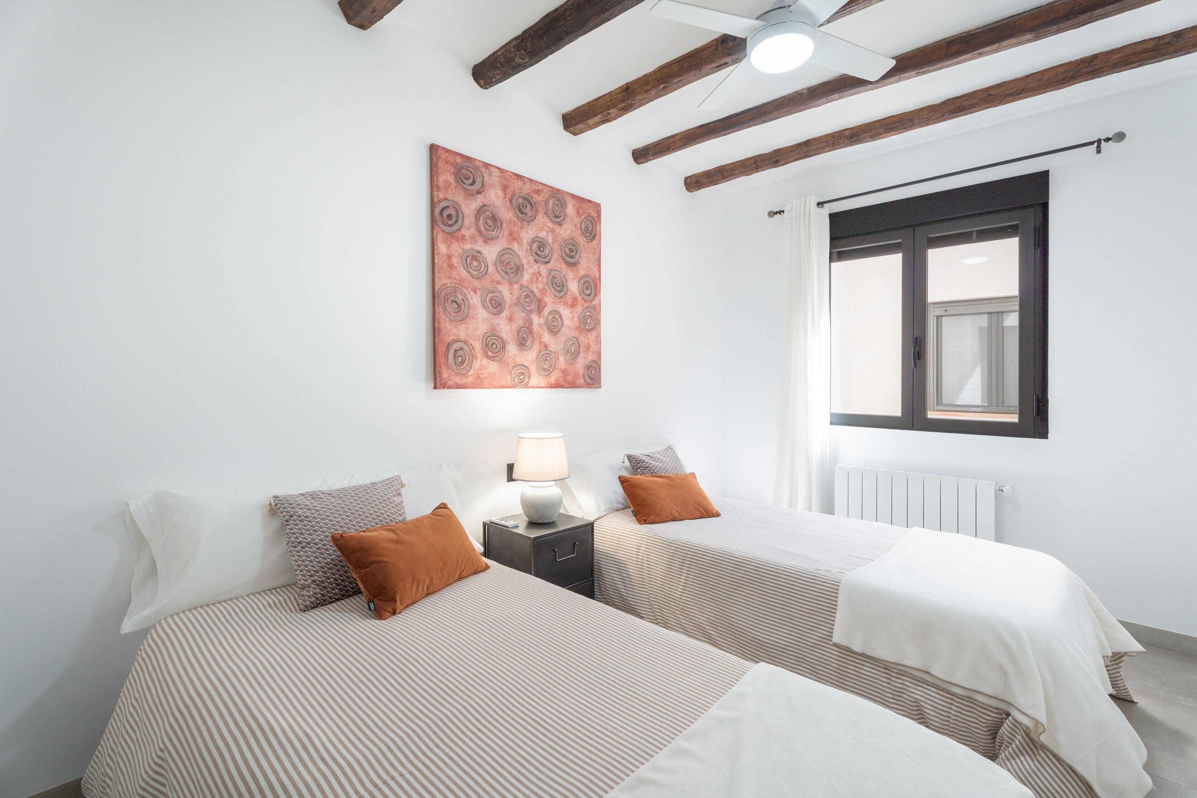 Camera d'albergo con due letti, una ventola a soffitto, una finestra con persiane e un'opera d'arte appesa al muro.
