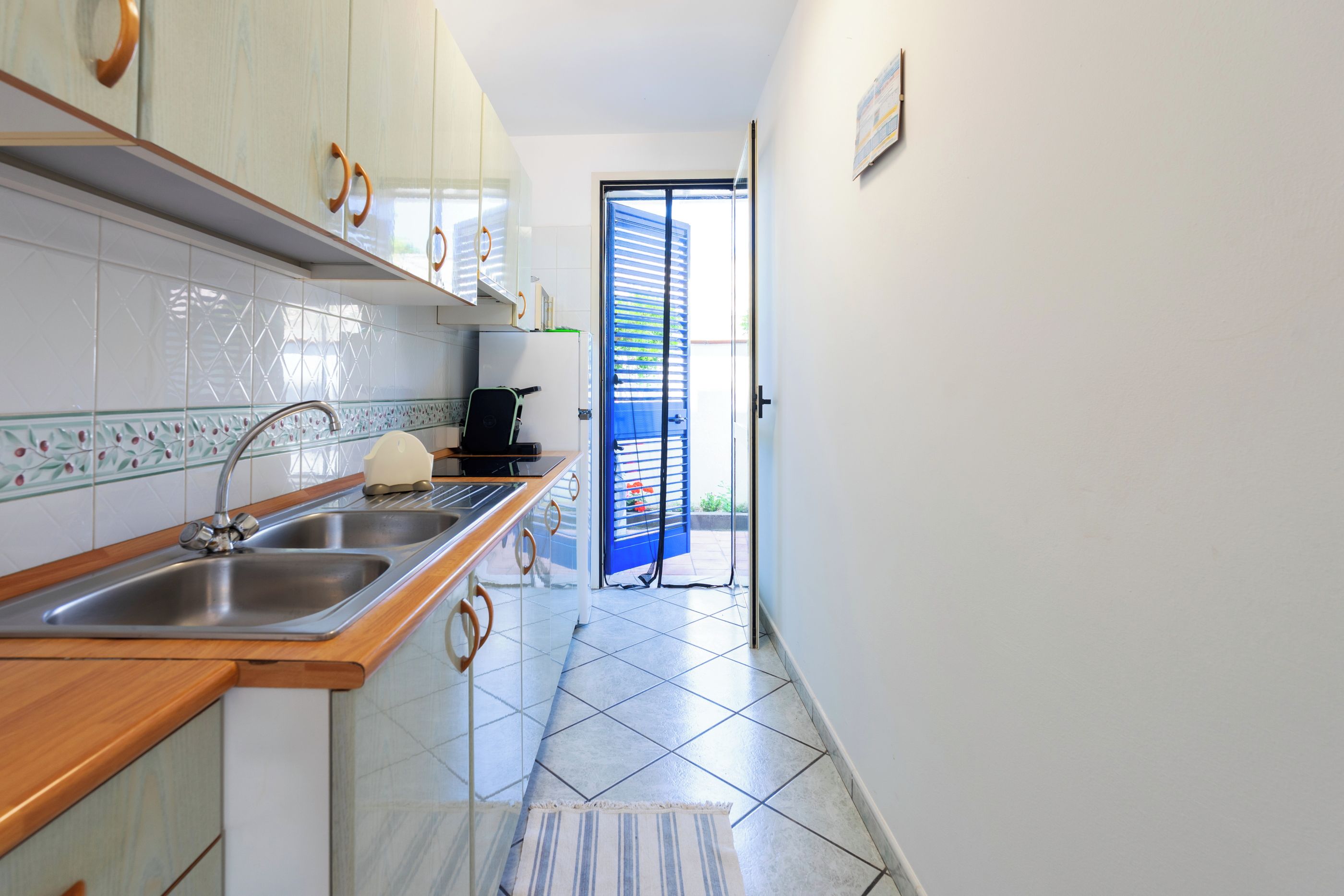 A narrow kitchen with a stainless steel sink, a kettle, and a toaster. There is a window with blinds, and a door leading outside.
