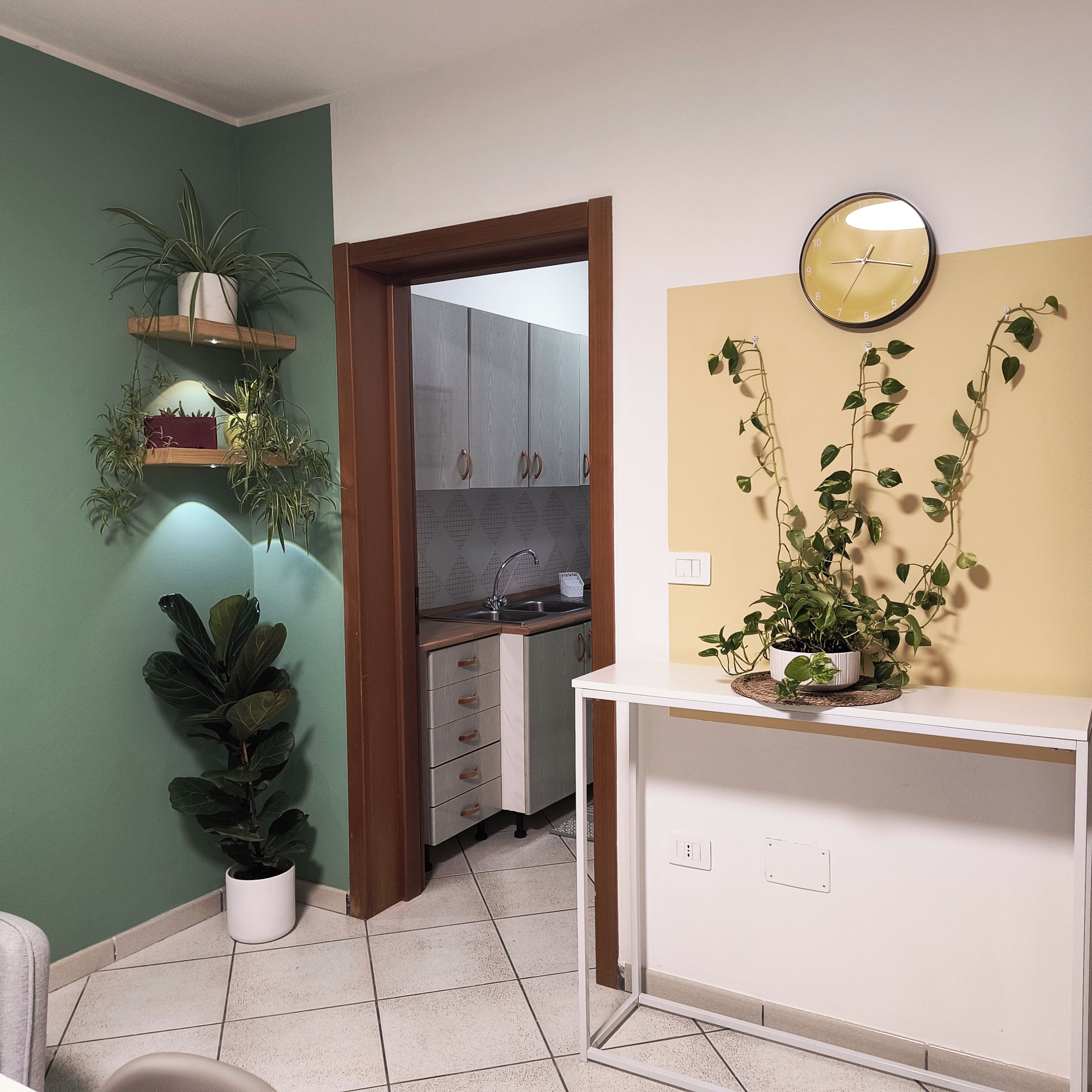 Una habitación con kitchenette, una mesita blanca con una planta en maceta y un reloj de pared.