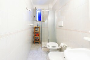 A bathroom with a toilet, a glass shower enclosure, and a small wooden shelf with a basket. - Residence Mare Azzurro (Ugento)