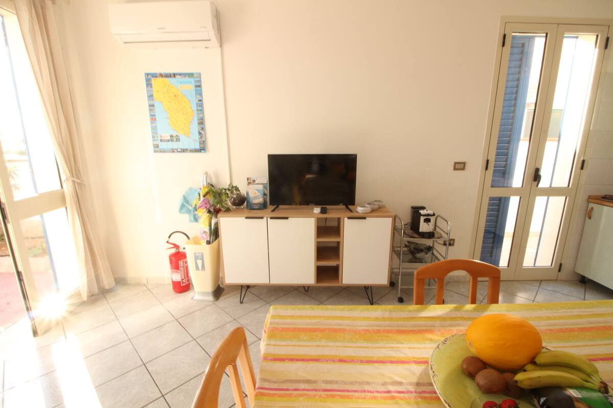 A room with a tiled floor, a table with a striped tablecloth, a television on a wooden cabinet, and a window with blinds.