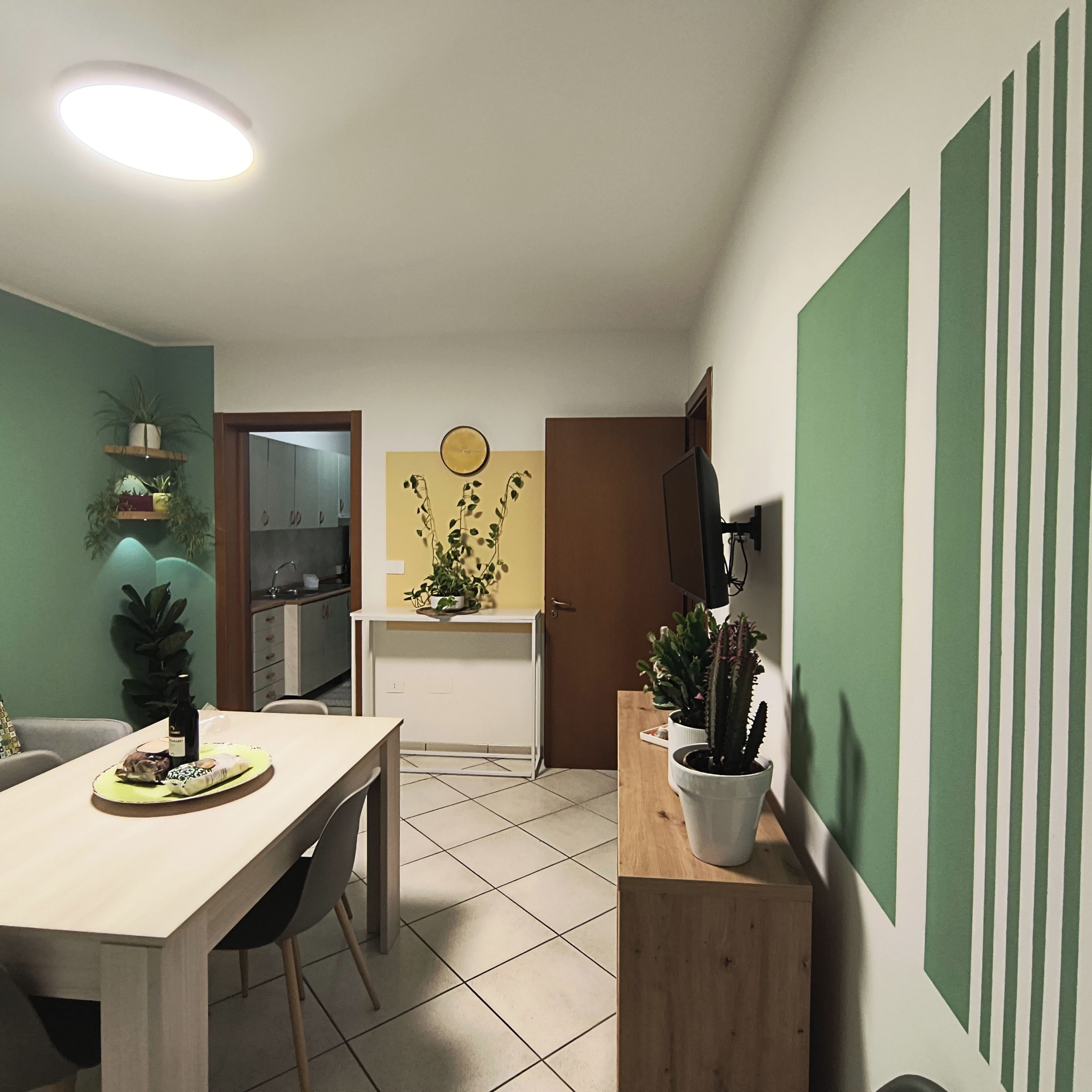 Comedor con una mesa y sillas, cocina al fondo y una pared verde con un reloj.