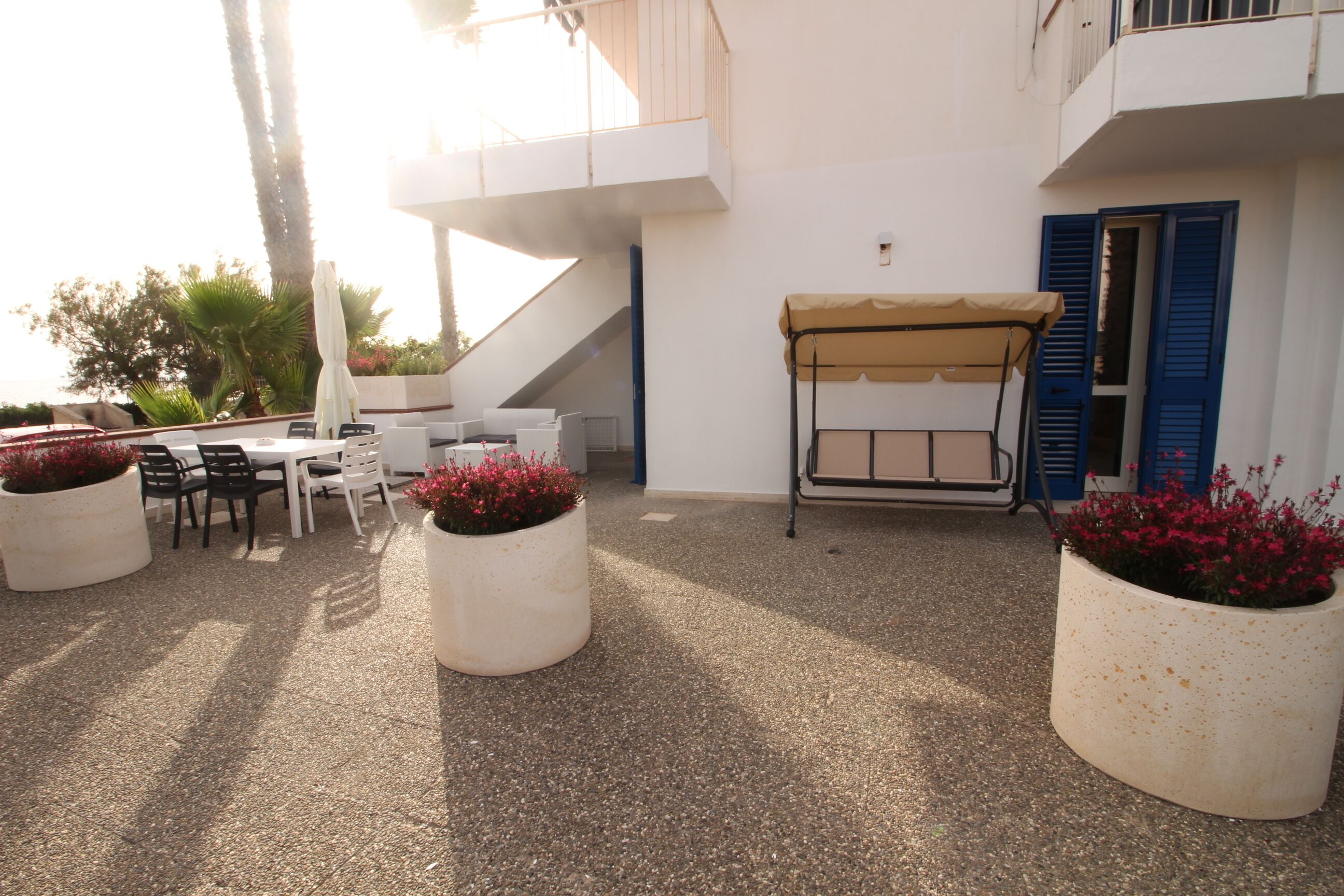 An outdoor patio with white tables and chairs, a covered seating area, and large potted plants with red flowers.