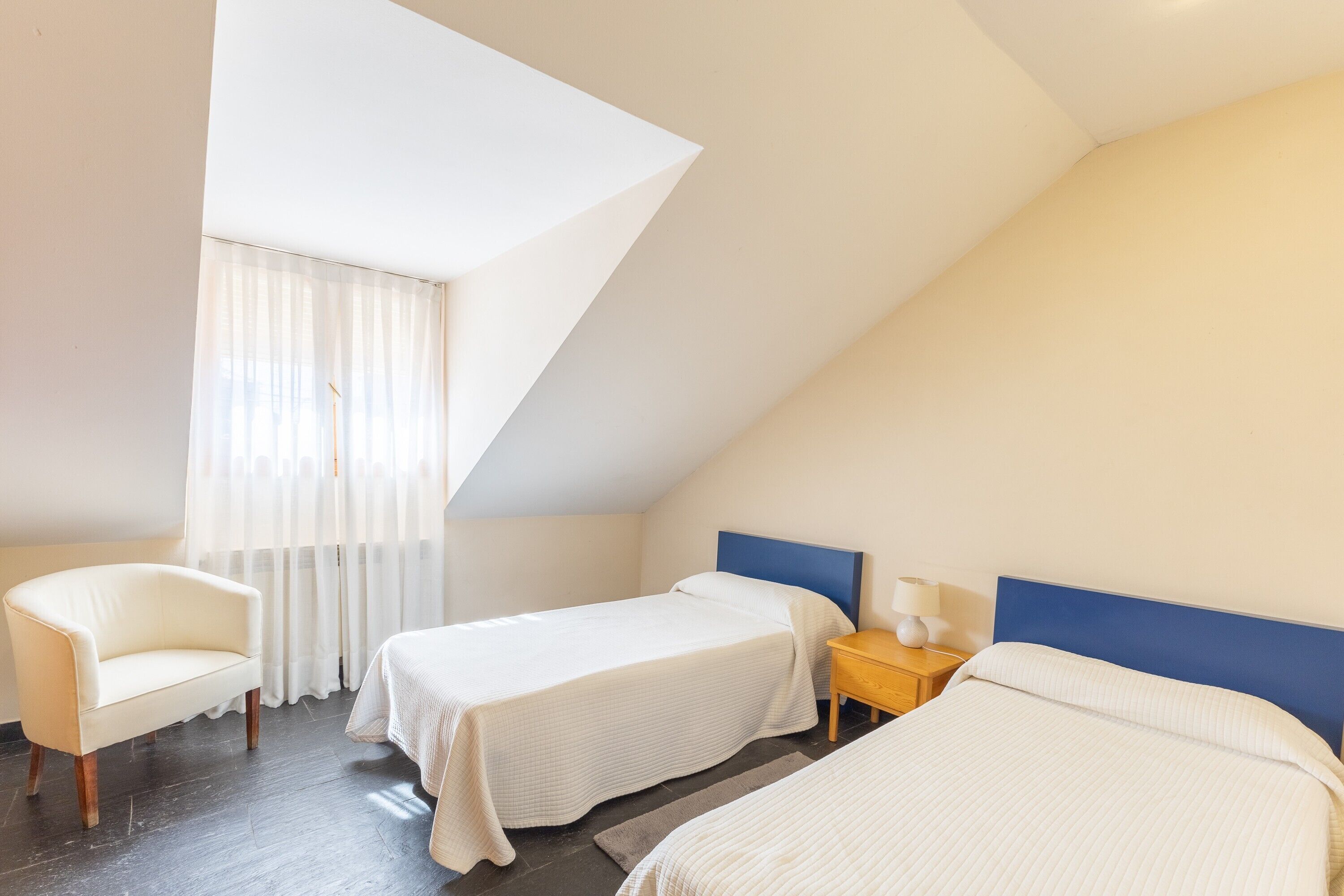 Una habitación con dos camas, una silla, una mesita de noche y una ventana con cortinas.