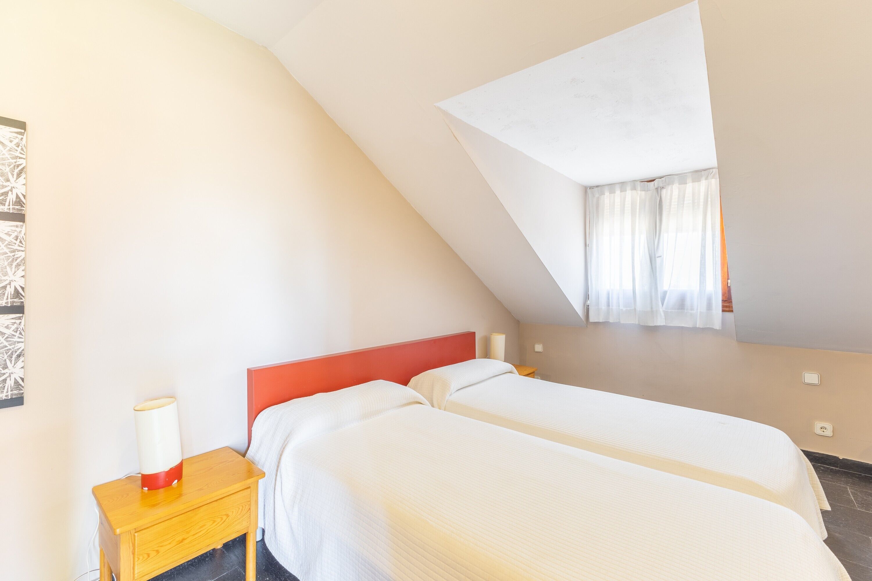A room with two beds, a nightstand, a lamp, and a window with curtains.