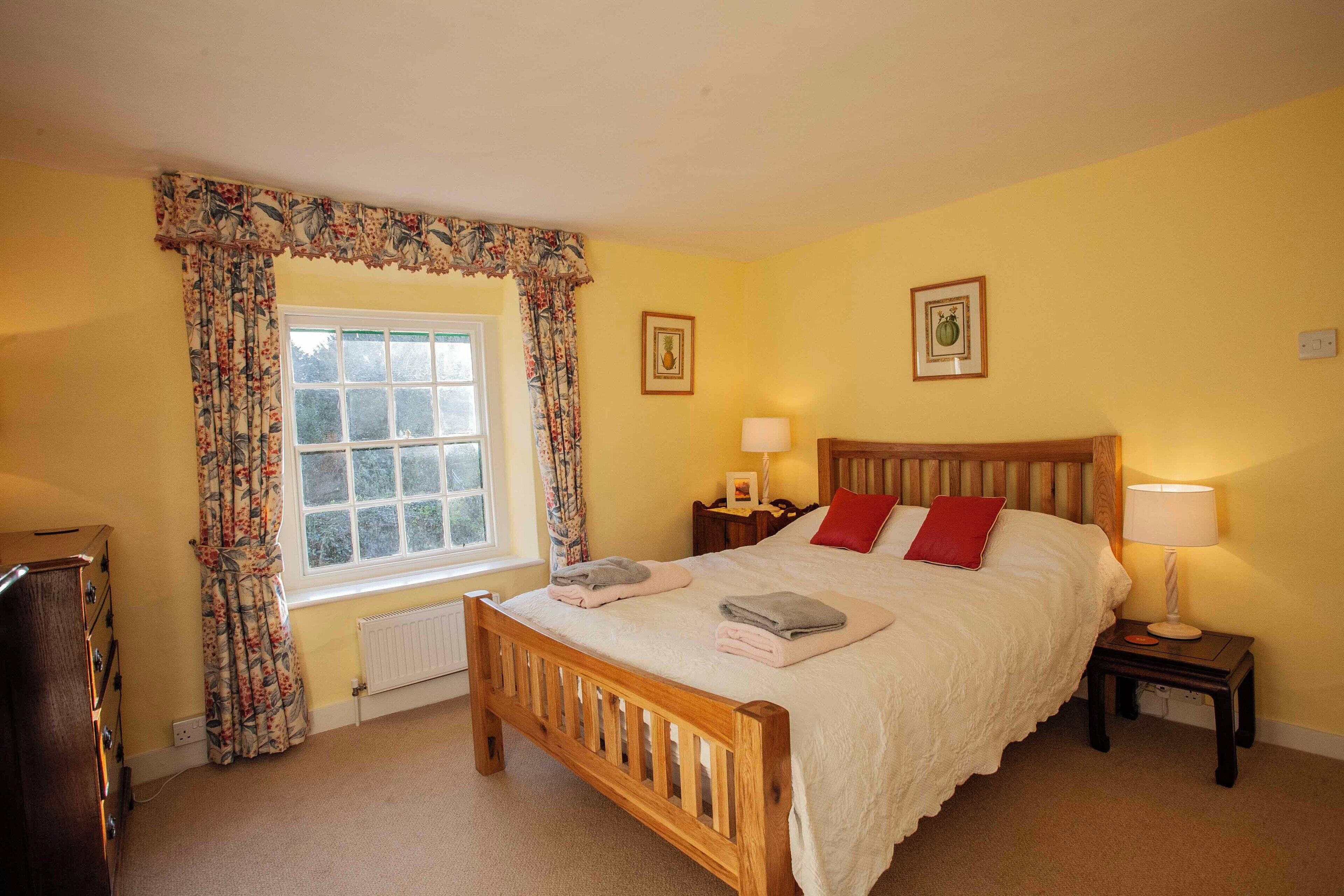 A bedroom with a wooden bed, two bedside tables with lamps, a window with floral curtains, and a radiator.