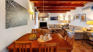 Un coin repas avec une table en bois, des chaises et un panier de fruits. On aperçoit une télévision au fond, un canapé et une lampe sur une table d’appoint.