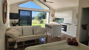 A modern, open-plan living and kitchen area with a large window, a ceiling fan, a sofa, a bed, and a kitchen with white cabinets and a stove. - Private Studio Byron Hinterland (Fernleigh)