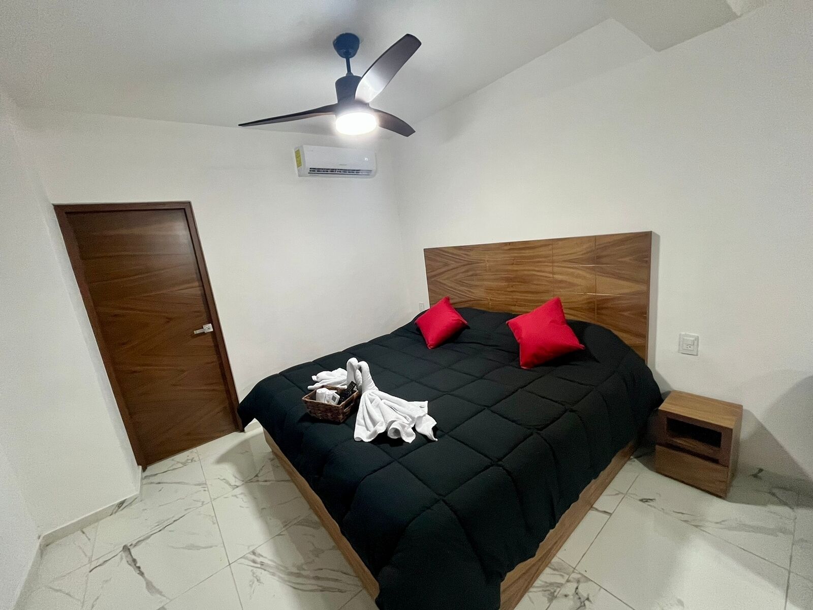 A bedroom with a bed, a ceiling fan, an air conditioner, and a wooden nightstand.