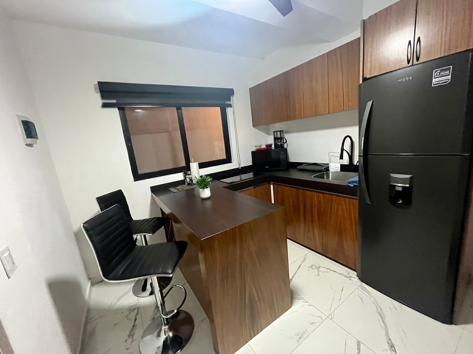 Cozinha compacta com armários de madeira, geladeira preta, cafeteira e uma pequena ilha com uma cadeira.