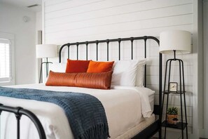 Un lit bien fait, recouvert d’une couverture bleue, avec des oreillers blancs et orange, et une tête de lit en métal noir. On y trouve deux lampes de chevet et une petite table sur laquelle sont posés une plante et une horloge.