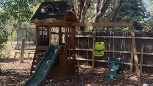 A wooden playhouse with a slide, surrounded by a fence and trees.