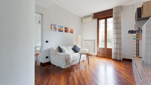 Ein Zimmer mit Holzboden, einem weißen Sofa mit Kissen, einem kleinen runden Tisch und einem Fenster mit Vorhängen.