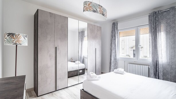 A modern hotel room with a large bed, a wardrobe, a desk, and a window with curtains.