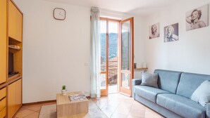 Uma sala de estar com sofá, mesa de centro, estante, relógio e porta que dá para uma varanda com vista.