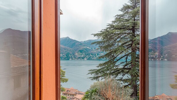 Vista pela janela de um lago, com montanhas ao fundo e um edifício com telhado vermelho em primeiro plano.