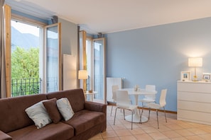 A room with a brown sofa, white chairs, a small table, a lamp, and a view of greenery through large windows.