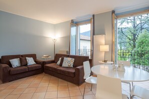 A living room with two brown sofas, a white table, and chairs, a floor lamp, and a view of greenery through large windows. - GuestHost - Cozy 60m2 apartment, perfect for two people, located on the 1st floor of a building with lift.The property is located on the road that connects Como and Menaggio, a few steps from the town center and just 300 meters from the Ossuccio lido, a  (Tremezzina)