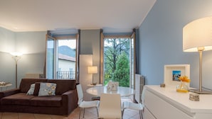 Un salon avec un canapé marron, des fauteuils blancs, une table ronde et une fenêtre donnant sur des arbres et des montagnes.