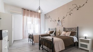 Un dormitorio con dos camas, una mesita de noche con lámpara, un armario y una ventana con cortinas translúcidas.
