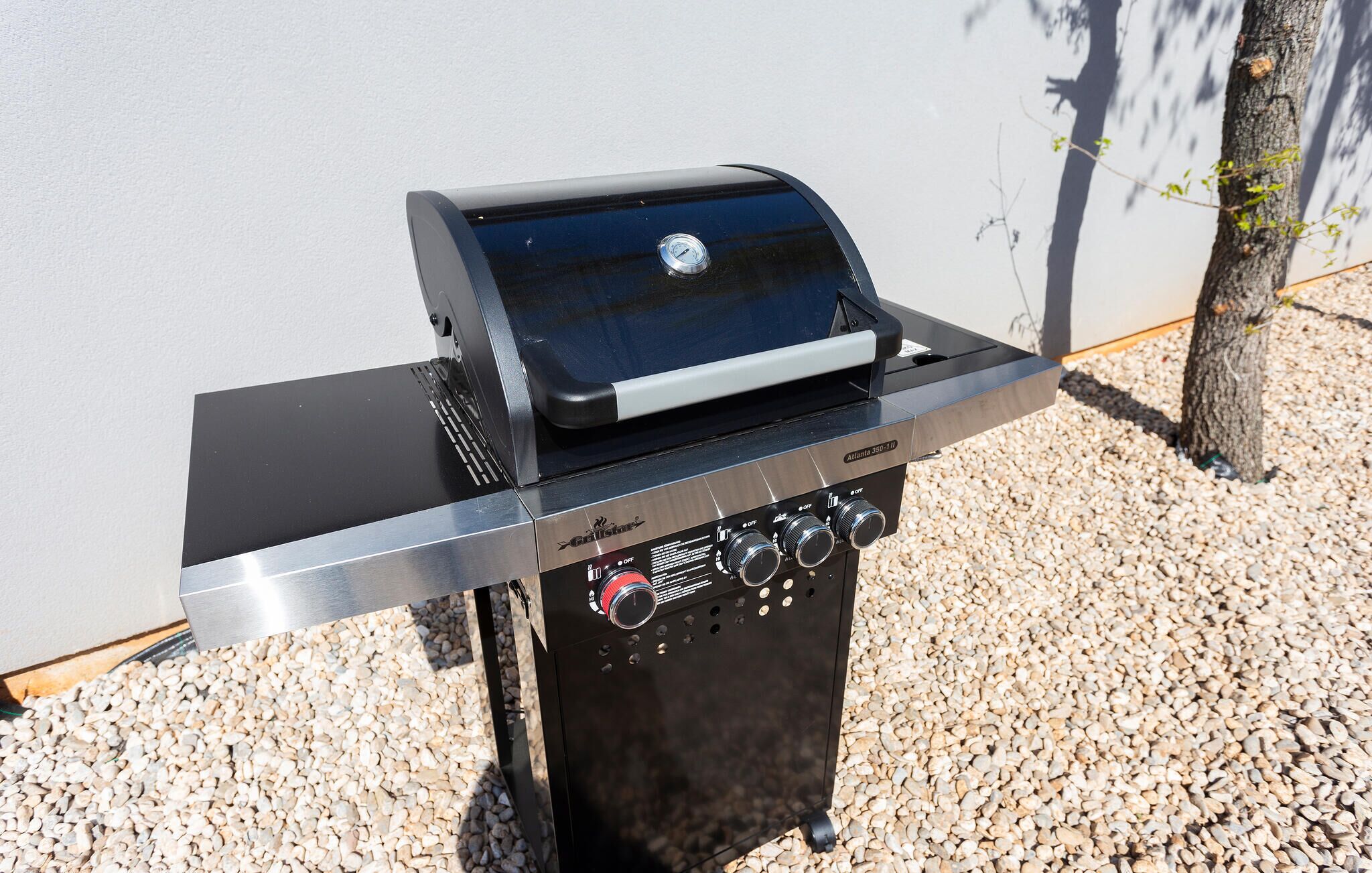 A portable grill with a digital display and control knobs, placed on a gravel surface with a tree in the background.