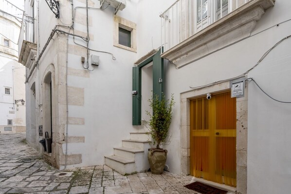 A narrow street with a building featuring a wooden door, a potted plant, and a staircase leading to an upper level. - Cozy studio apartment that comfortably accommodates two people, equipped with all the main comforts for a pleasant stay. The location is ideal: the accommodation is in fact located in the center (ZTL), with all the essential services within reach and a v (Martina Franca)