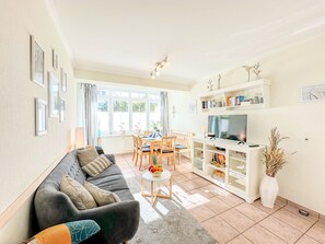 Ein Wohnzimmer mit einer Couch, einem Esstisch, Stühlen, einem Fernseher und einem Bücherregal.