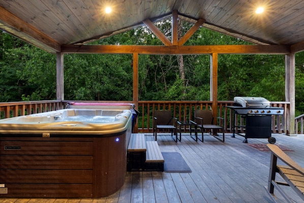 Terraza exterior techada con jacuzzi, sillas, una parrilla y barandilla de madera.