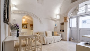 A room with a white sofa, a wooden chair, a small table, and a kitchen area with a white cabinet and a window. - GuestHost - White Lotus Apartment (Ostuni)