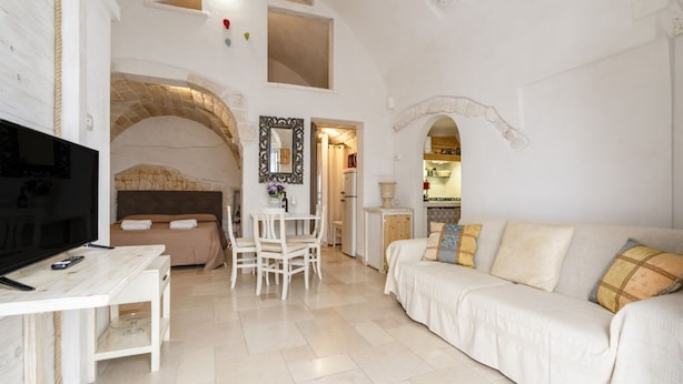 A spacious living area with a TV, a sofa, a dining table, and a kitchen in the background. - GuestHost - White Lotus Apartment (Ostuni)