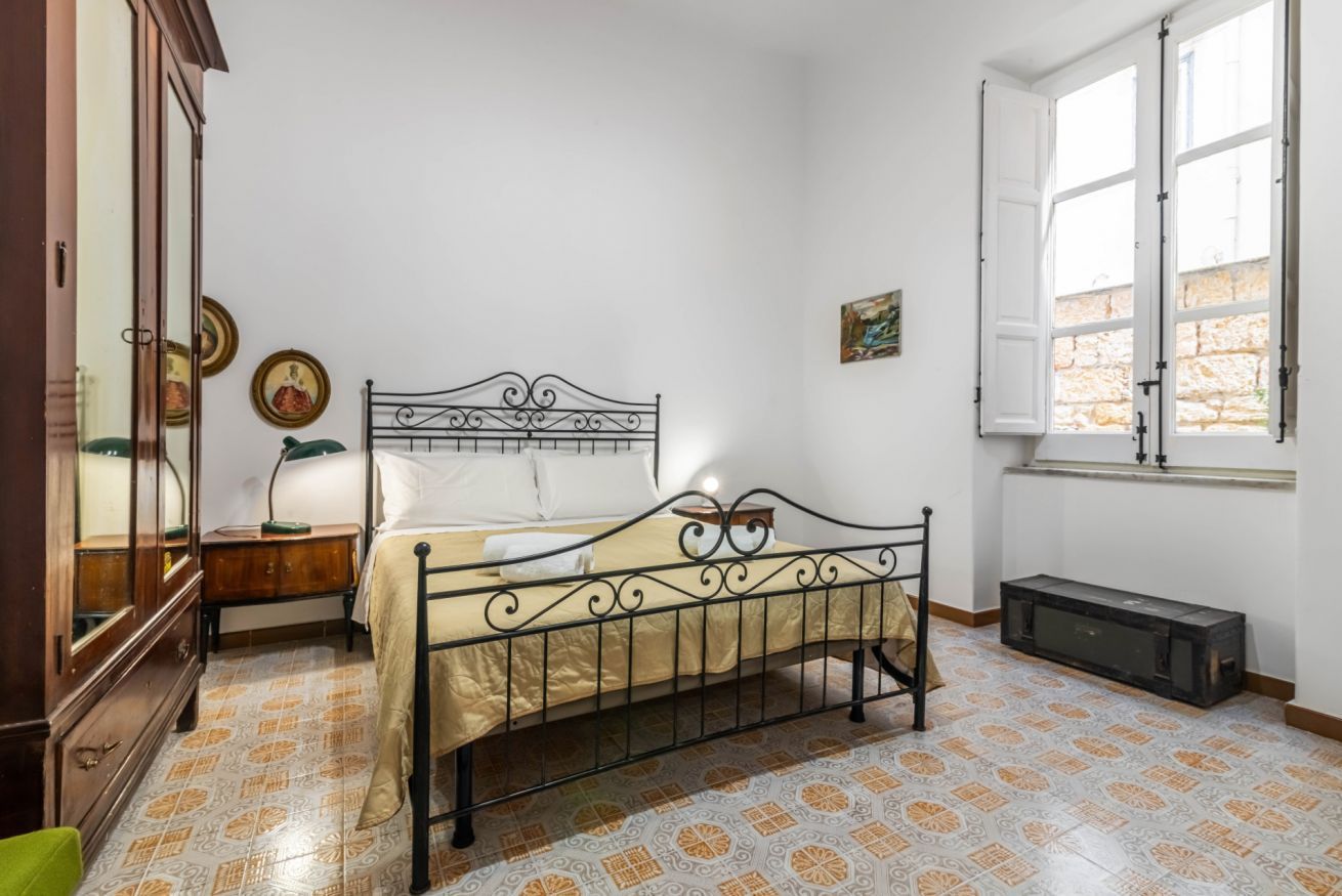 A bedroom with a metal bed frame, a wooden dresser, a small bedside table, a window with white shutters, and patterned floor tiles.