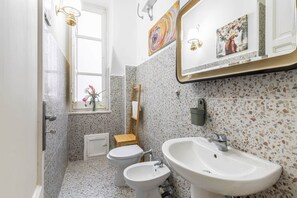 Een badkamer met toilet, bidet en wastafel, betegeld met een streeppatroon, en een raam met uitzicht op bloemen.