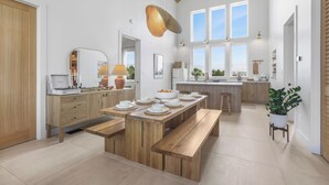 A spacious kitchen with a wooden dining table set for six, a large island, and a view of the outdoors through the windows.