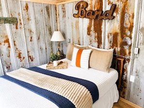 A rustic bedroom with a bed, pillows, a lamp, and a wall-mounted sign.