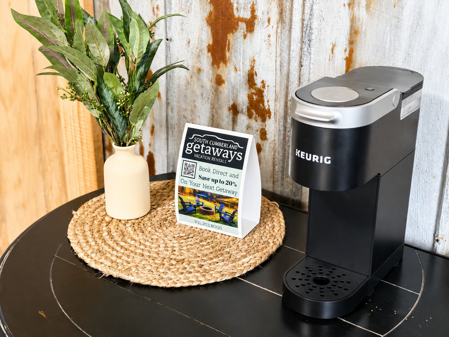 A KEURIG coffee maker on a black countertop with a woven placemat, a sign for South Cumberland getaways, and a potted plant.