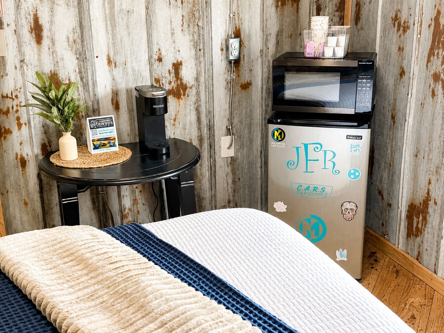 A room with a bed, a small table with a Keurig coffee maker, a microwave, and a refrigerator with stickers.