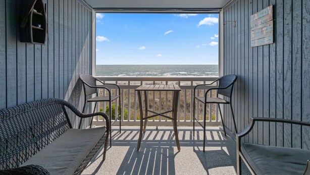 A balcony with a view of the ocean, a wooden table, two chairs, and a wicker sofa. - Windy Shores-Oceanfront 2BD Condo-Pool-Pet Friendly (North Myrtle Beach)