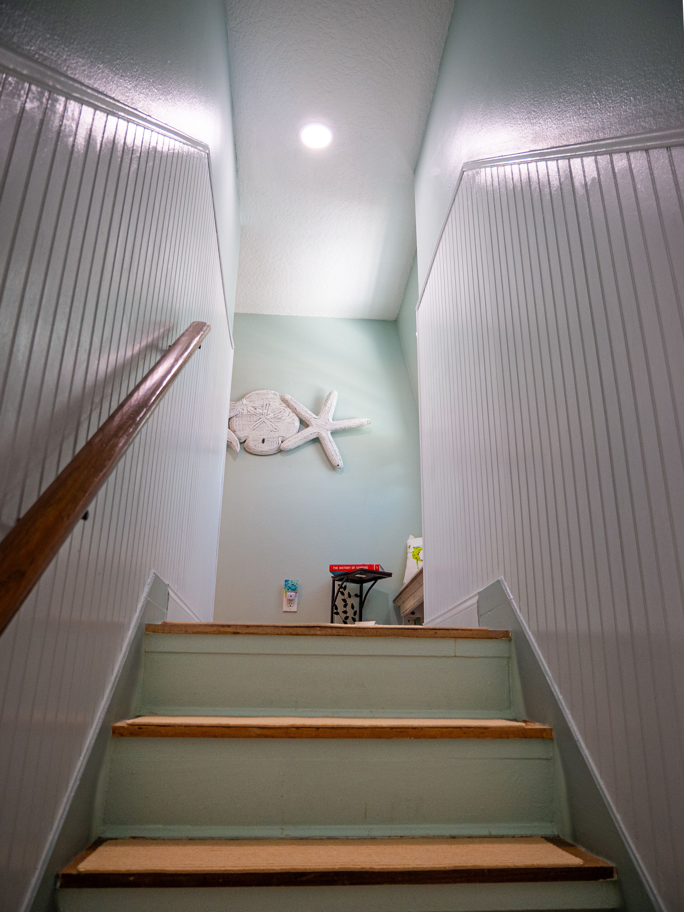 Un escalier avec des marches en bois et une rampe, menant à un palier orné d’une étoile de mer fixée au mur et sur lequel se trouve une petite table avec un livre.