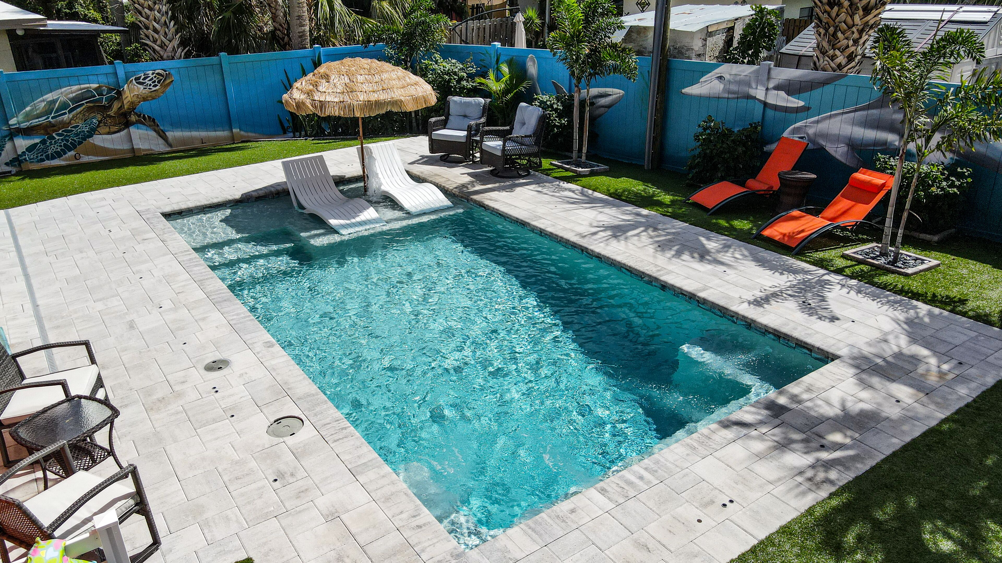 Une piscine rectangulaire à l’eau d’un bleu limpide, entourée d’une terrasse carrelée. On y trouve des chaises longues et un parasol en chaume.
