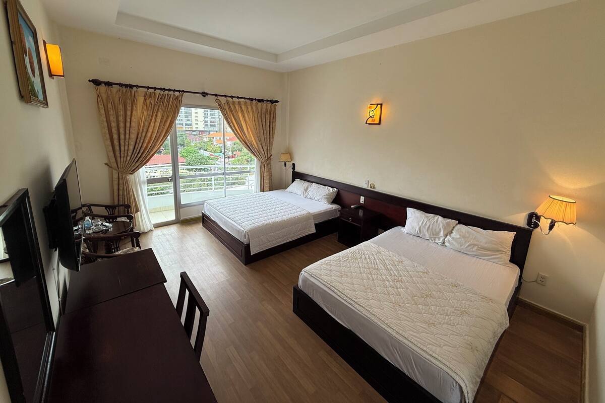 A hotel room with two beds, a desk, and a chair. There is a window with curtains, a wall-mounted lamp, and a painting.