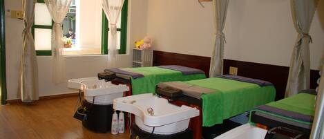 A room with three beds, each with a green bedspread and a headboard. There are two sinks with faucets, and a window with curtains.