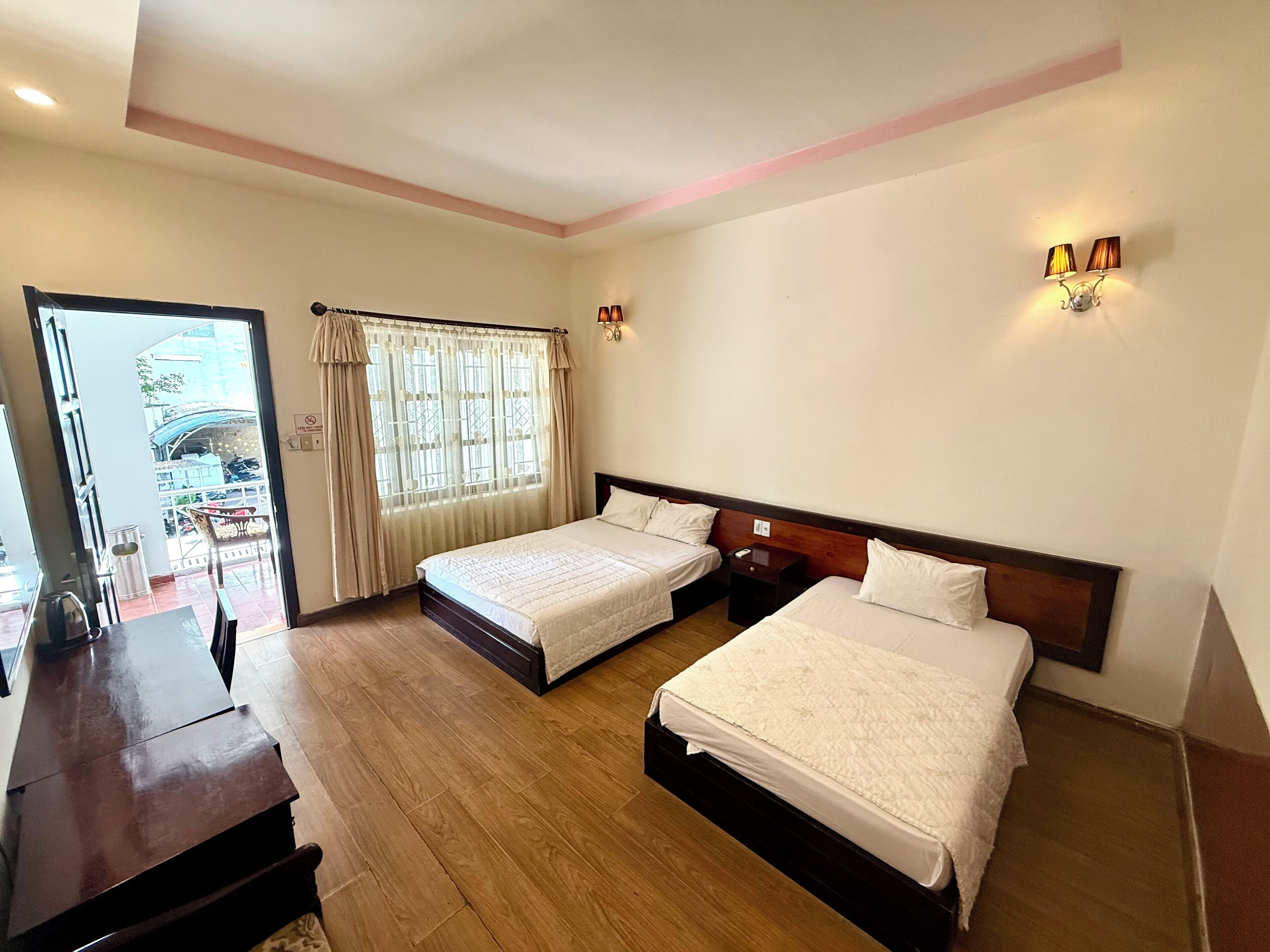 Une chambre d’hôtel avec deux lits, un bureau en bois, une fenêtre avec des rideaux et une lampe fixée au mur.