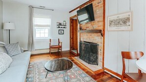 Een woonkamer met een bank, stoelen, een salontafel, een open haard, een aan een bakstenen muur bevestigde televisie en een raam met gordijnen.