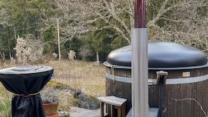 A hot tub with a wooden exterior and a metal chimney, surrounded by a wooden deck and potted plants.
